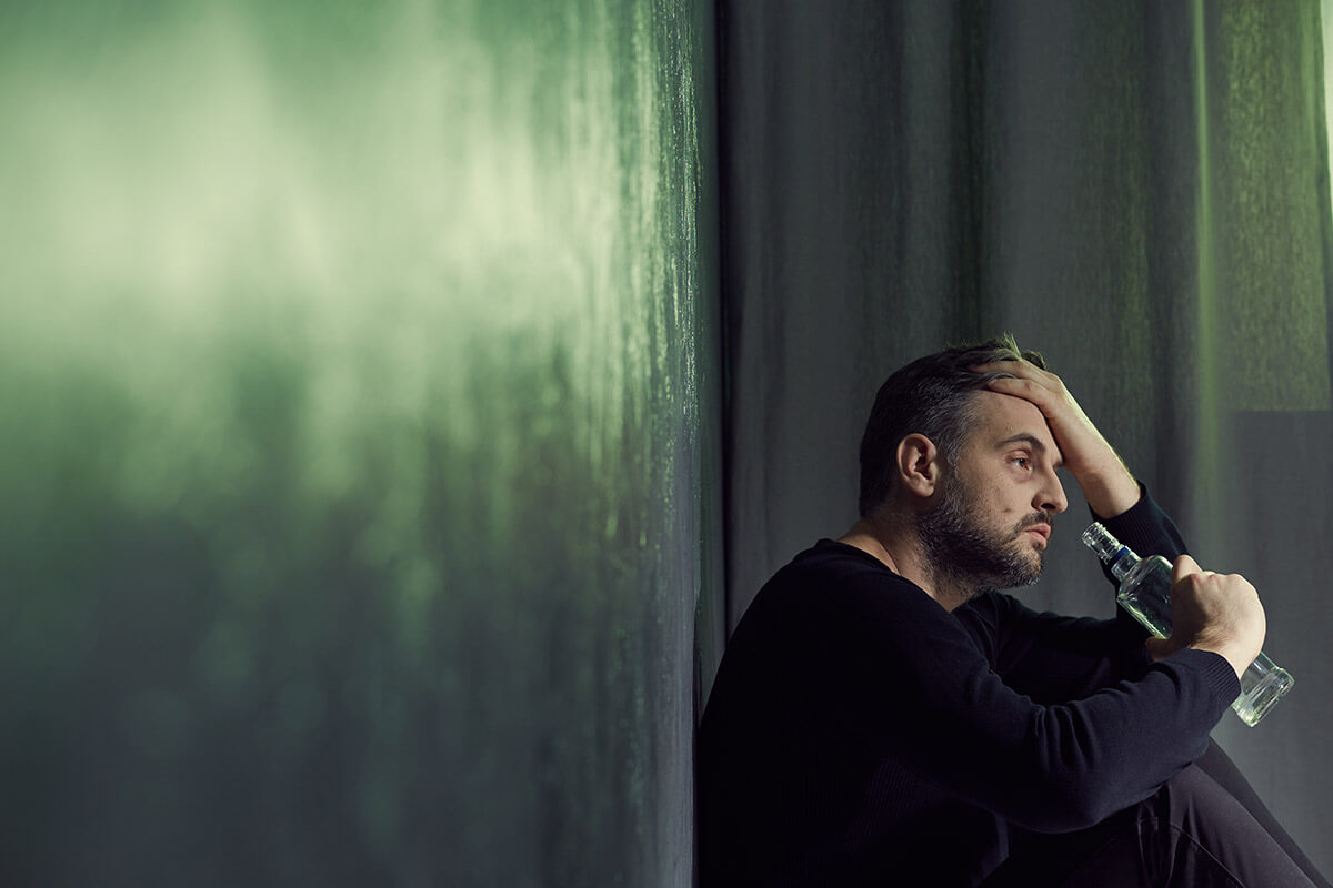 dangers of alcohol withdrawal man sitting against wall drinking alcohol to postpone the Dangers of Alcohol Withdrawal